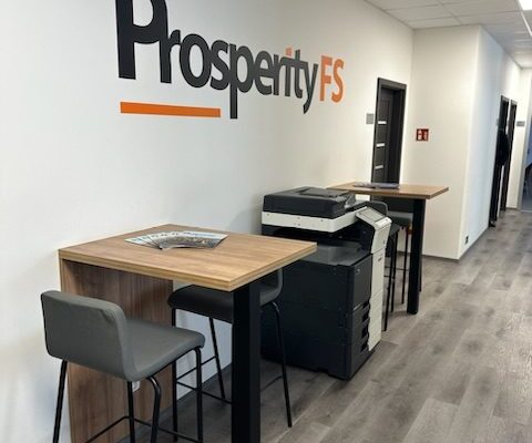 Interiér moderní kanceláře s logem Prosperity FS na stěně, vysokým stolkem s barovou židlí a tiskárnou.